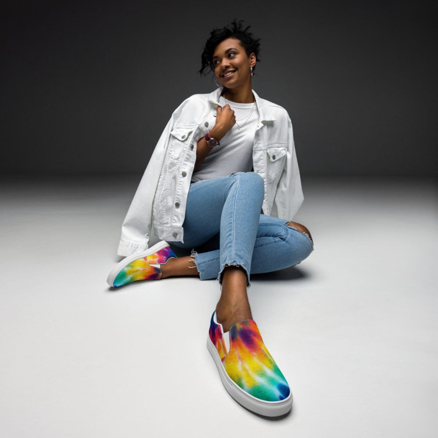 Stylish woman posing confidently in a studio setting wearing vibrant rainbow tie-dye slip-on sneakers, light wash ripped skinny jeans, a white t-shirt, and a white denim jacket draped over her shoulders. The bold multicolor canvas shoes feature a smooth blend of red, orange, yellow, green, blue, and purple hues with a clean white sole.