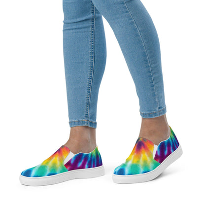 Close-up of a woman wearing rainbow tie-dye slip-on canvas sneakers with light blue skinny jeans on a white background. These vibrant shoes feature a bold, multicolor watercolor pattern in shades of blue, green, yellow, orange, red, and purple. Designed with a sleek white rubber sole and elastic side panels for easy on/off comfort.