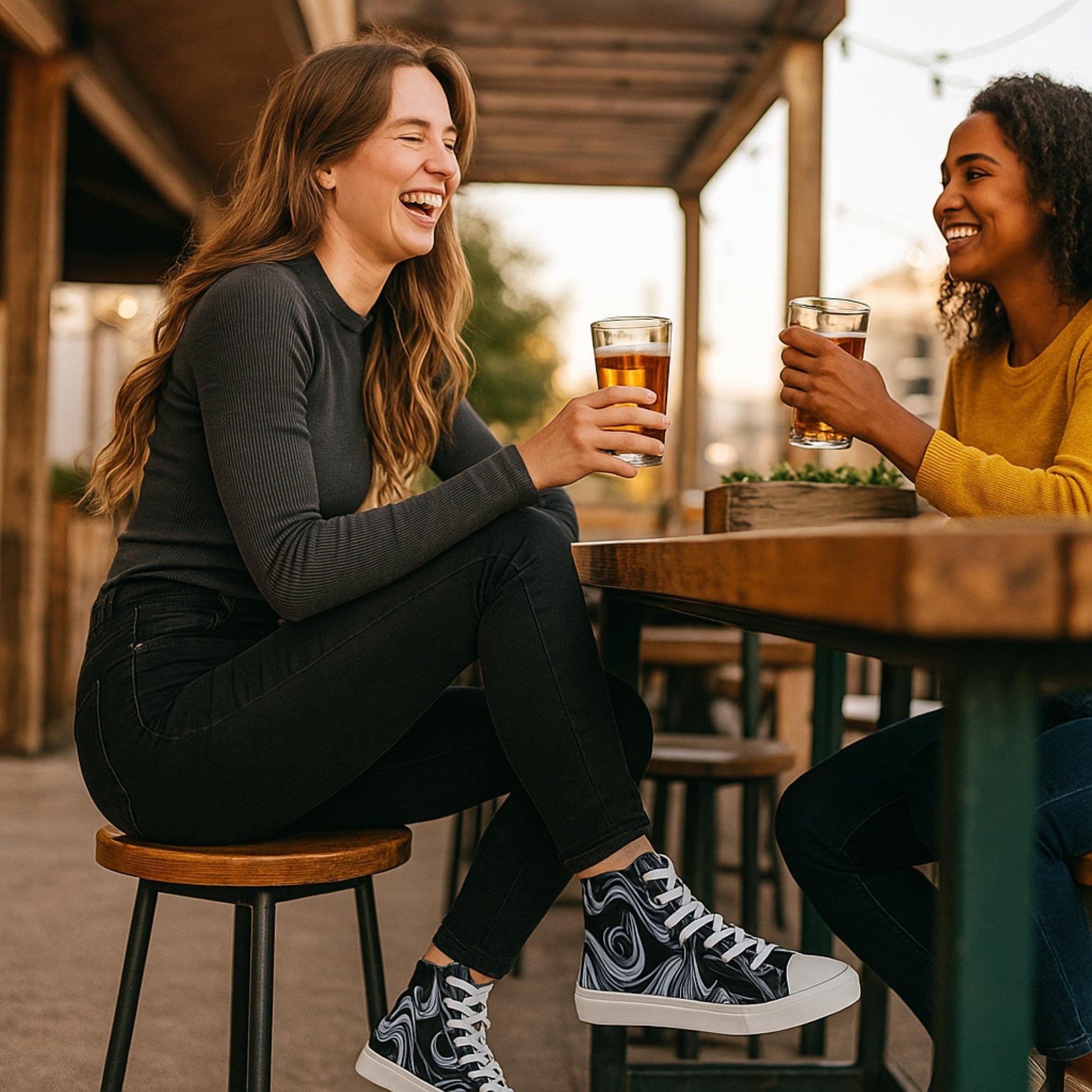 Two women are sitting at an outdoor wooden table, enjoying drinks and laughing together. The woman on the left is wearing a black ribbed long-sleeve top, black jeans, and high-top sneakers with a black and white abstract swirl pattern, white laces, and white toe caps. The setting has a casual and warm atmosphere with string lights and wooden structures in the background.