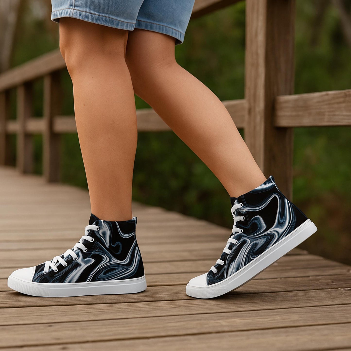 Person walking on a wooden bridge wearing high-top sneakers with a bold black, white, and blue marble swirl pattern, white laces, and white soles. The person is dressed in light denim shorts, and the background features blurred greenery, suggesting a nature trail or park setting.