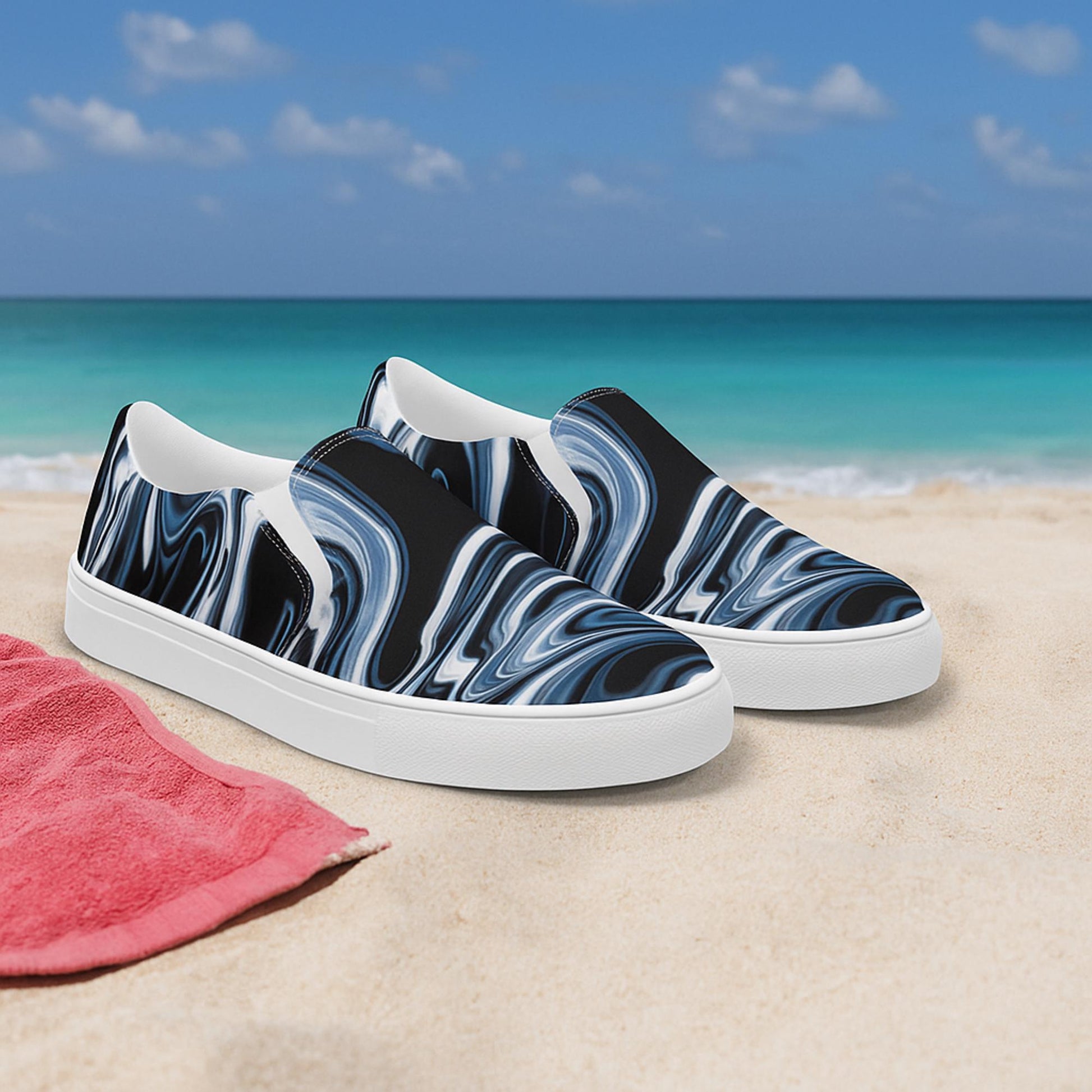 Pair of women’s slip-on canvas shoes with a black, white, and gray marbled swirl design displayed on sandy beach with turquoise ocean and blue sky in the background. Stylish and comfortable beachwear shoes, ideal for summer fashion.