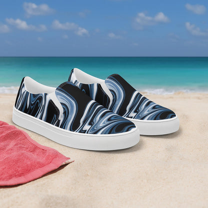 Pair of women’s slip-on canvas shoes with a black, white, and gray marbled swirl design displayed on sandy beach with turquoise ocean and blue sky in the background. Stylish and comfortable beachwear shoes, ideal for summer fashion.