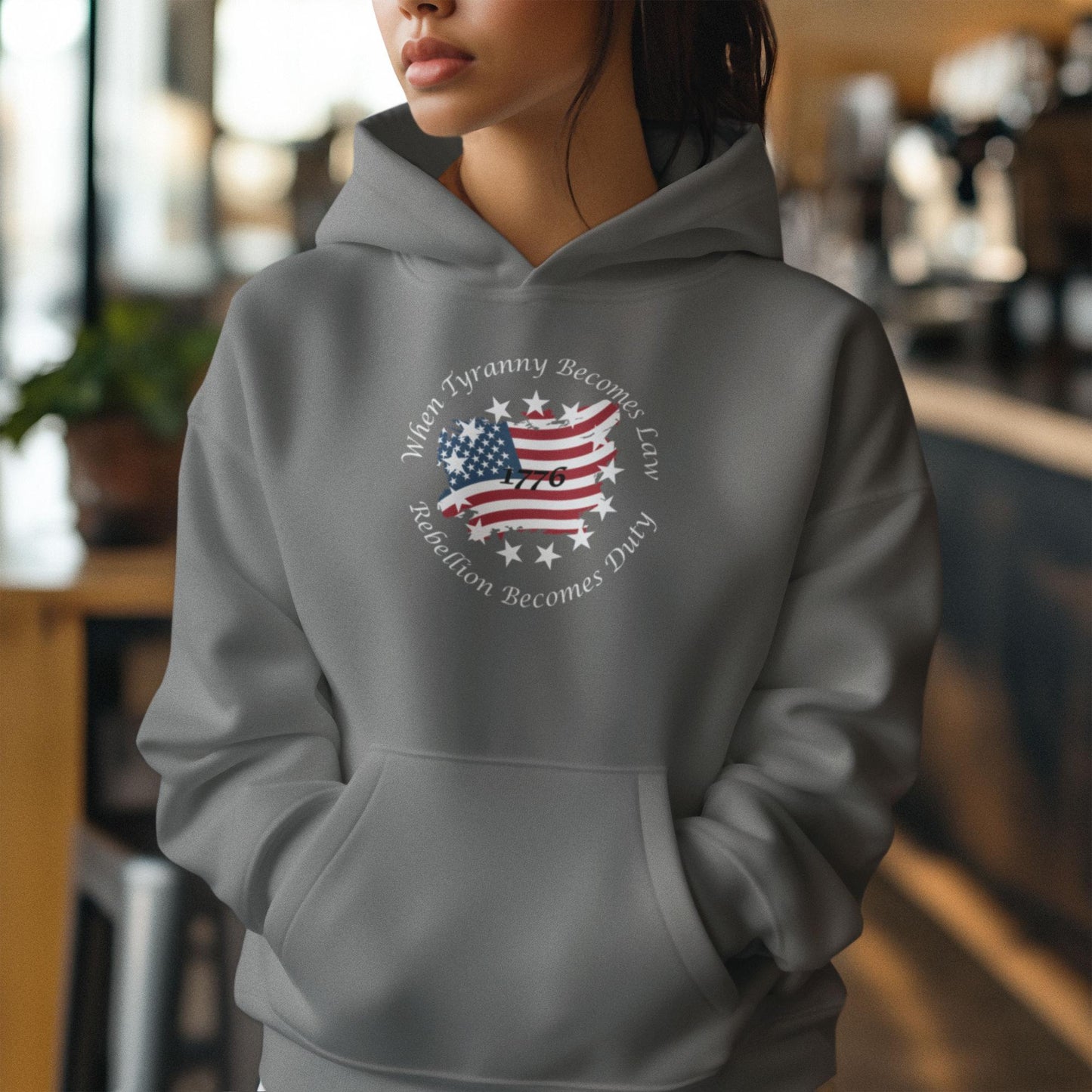 Gray patriotic hoodie with distressed American flag and “When Tyranny Becomes Law, Rebellion Becomes Duty” quote, worn by woman indoors. 1776 graphic sweatshirt for women, conservative USA freedom apparel, revolutionary slogan pullover, perfect for pro-America fashion, political activism wear, or patriotic gift for her.