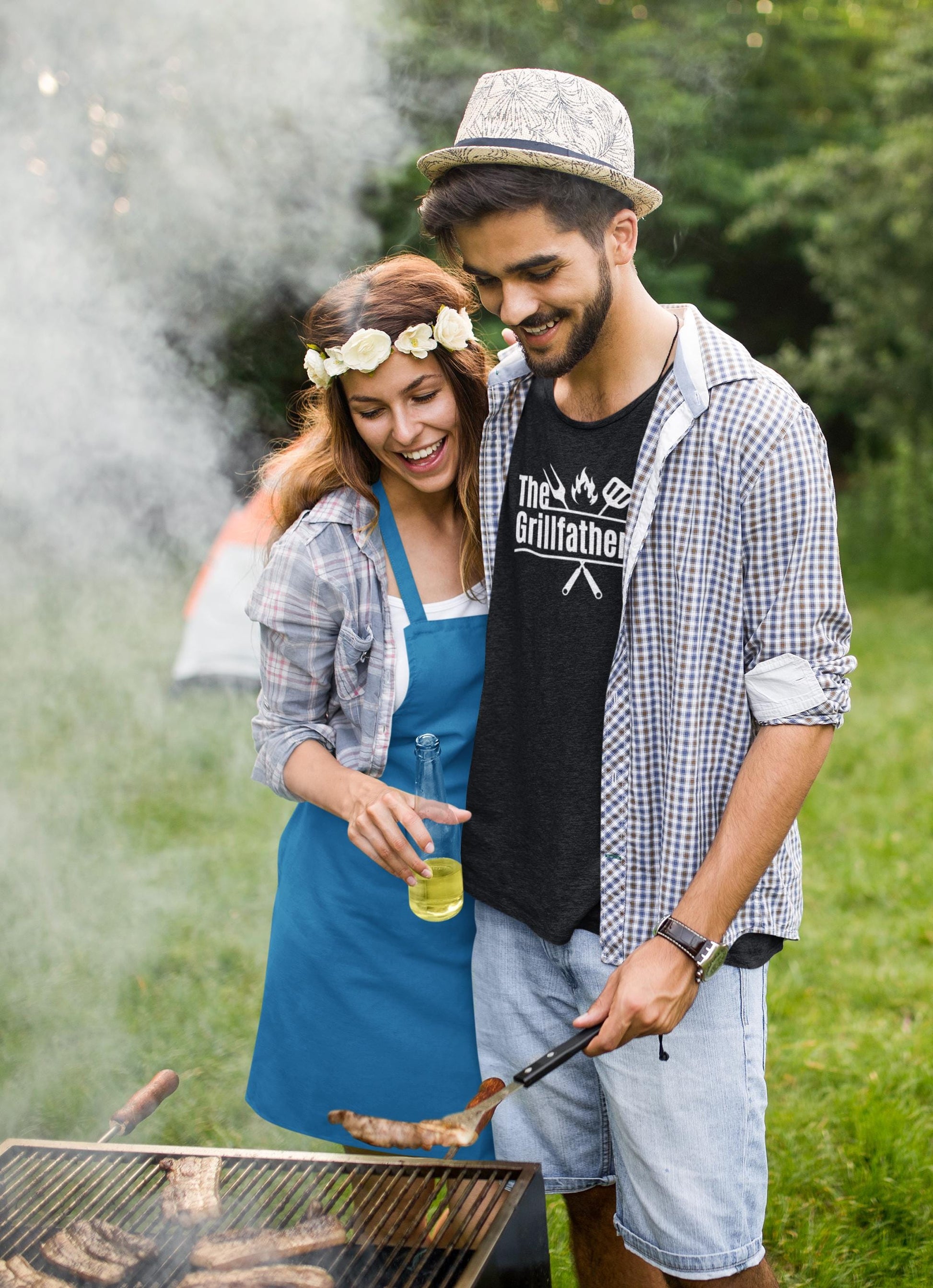 Man wearing a black T-shirt with the phrase “The Grillfather” and grilling icons, smiling while barbecuing outdoors with a woman in a flower crown and blue apron holding a drink.