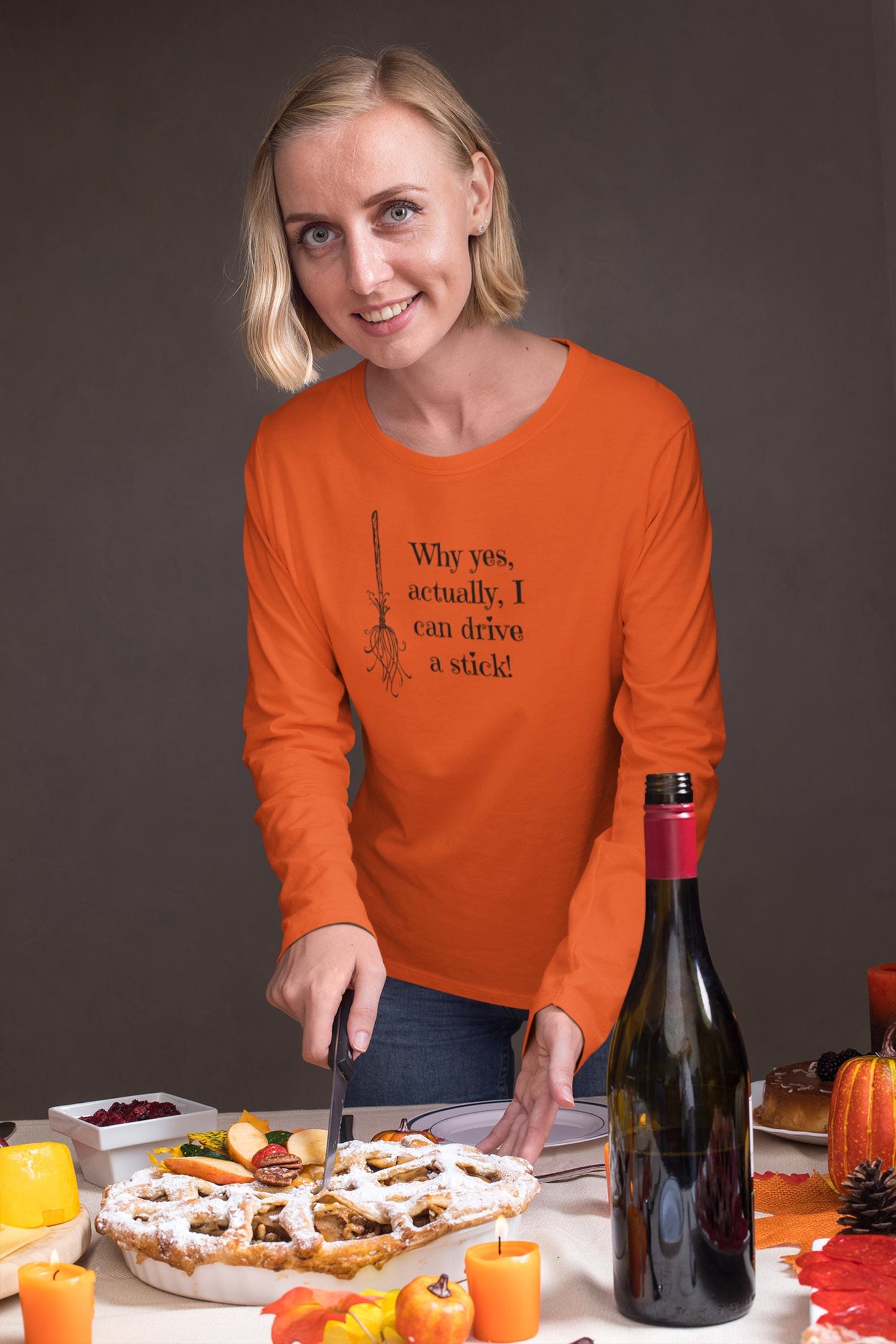 Woman wearing a festive orange long-sleeve Halloween shirt with a black broomstick graphic and the funny witch-themed saying &quot;Why yes, actually, I can drive a stick!&quot; — perfect for spooky season parties and fall celebrations. Great Halloween apparel for women, humorous witch shirt for autumn gatherings.