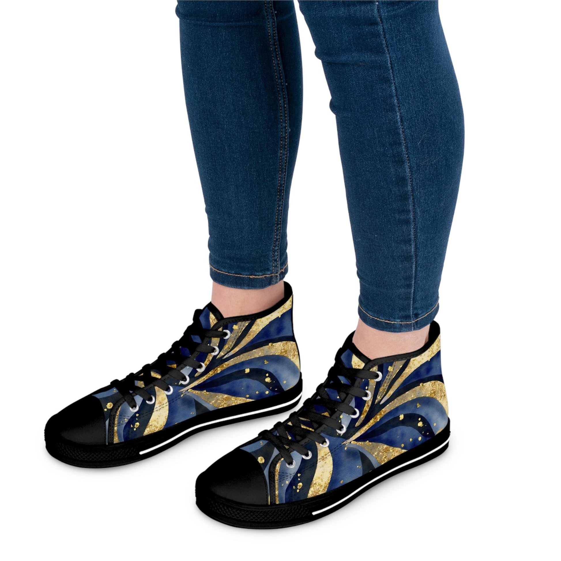 Close-up view of a person wearing high-top sneakers with a vibrant abstract design in navy blue, gold, and black, featuring swirling shapes and dotted accents. The shoes have black laces, black rubber soles, and black toe caps, paired with dark blue skinny jeans. Set against a plain white background.