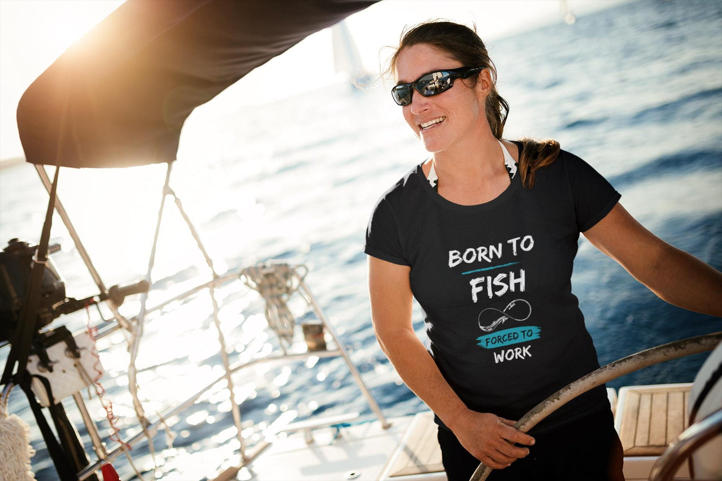 Smiling woman steering a sailboat on the open water, wearing sunglasses and a black t-shirt with a graphic that reads &#39;Born to Fish, Forced to Work.&#39; The design includes a fishing hook illustration and features white and teal text. The sun is shining brightly, creating a warm and adventurous atmosphere.