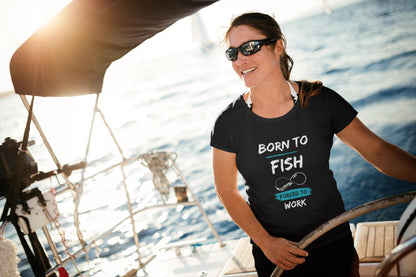 Smiling woman steering a sailboat on the open water, wearing sunglasses and a black t-shirt with a graphic that reads &#39;Born to Fish, Forced to Work.&#39; The design includes a fishing hook illustration and features white and teal text. The sun is shining brightly, creating a warm and adventurous atmosphere.