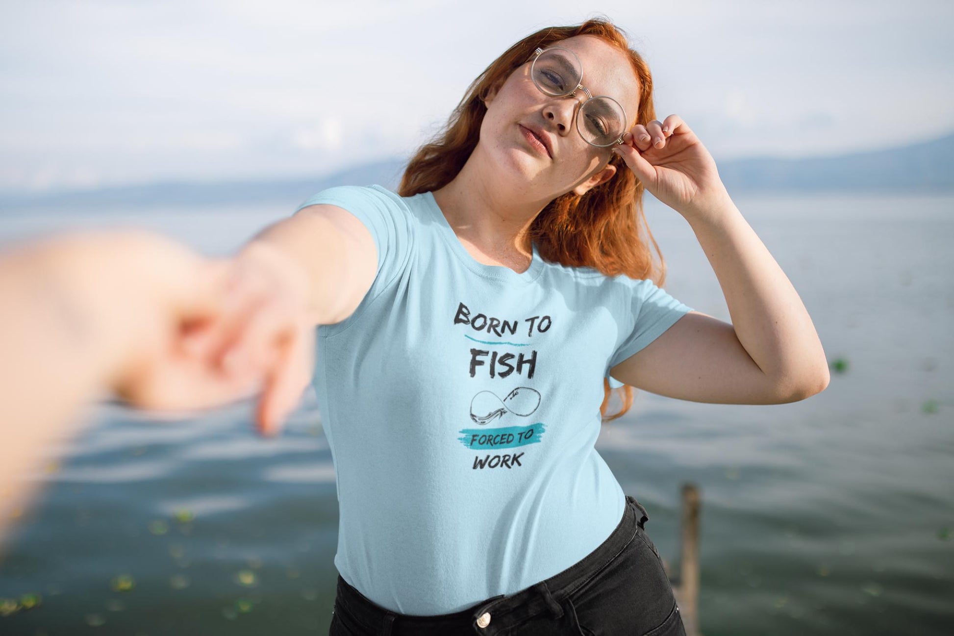 Confident woman standing by a lake, holding someone&#39;s hand and wearing a light blue t-shirt with the phrase &#39;Born to Fish, Forced to Work&#39; in black and teal text. The design includes a simple fishing hook graphic. She has long red hair, is wearing glasses, and poses with a relaxed, playful expression.