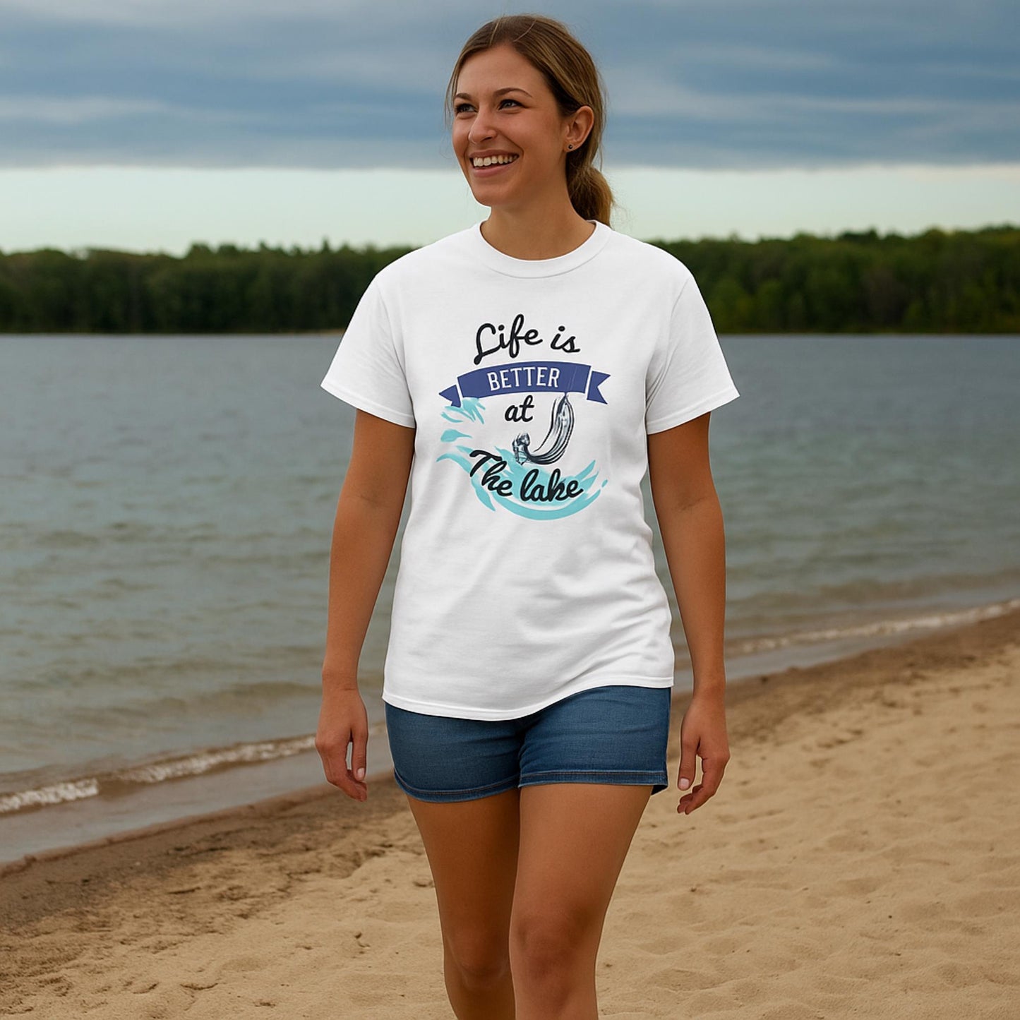 Smiling woman walking along a sandy lakeshore, wearing a white t-shirt with a colorful graphic that reads &#39;Life is Better at the Lake&#39; featuring a fishing hook, waves, and banner design. She pairs the shirt with denim shorts and is surrounded by a scenic lake and forested background under a cloudy sky.