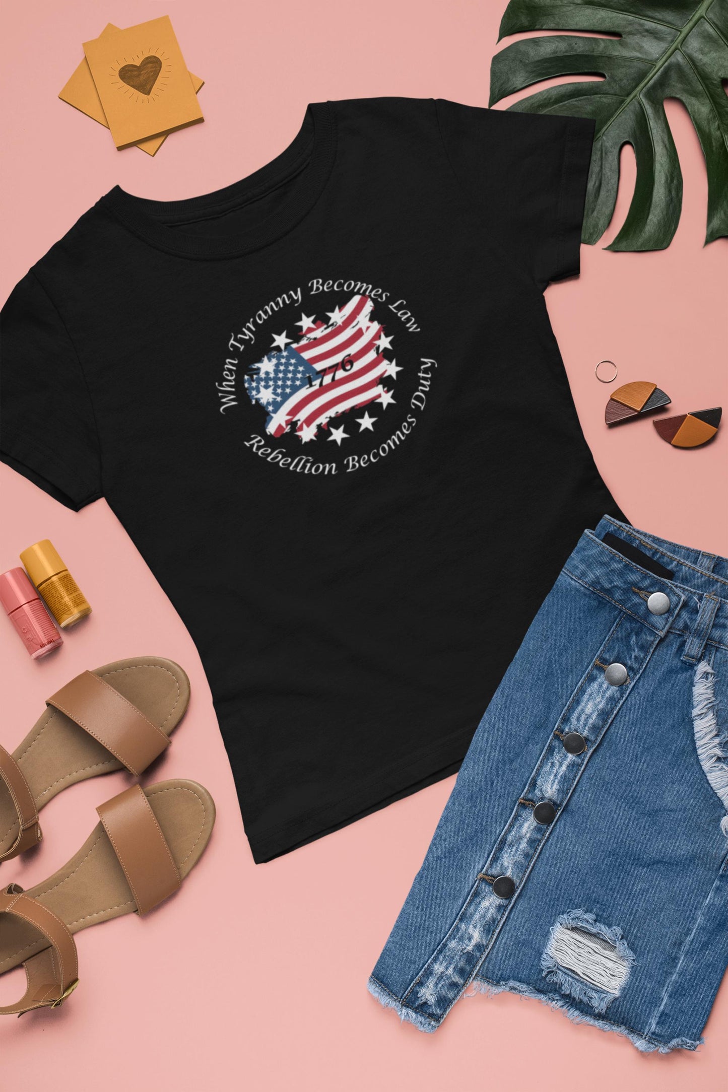 Flat lay of a black women&#39;s fitted t-shirt with a patriotic design featuring a distressed American flag shaped like the U.S., surrounded by stars and the quote &#39;When Tyranny Becomes Law, Rebellion Becomes Duty - 1776.&#39; Styled alongside a ripped denim skirt, tan sandals, earrings, lip gloss, and a tropical leaf on a pink background.