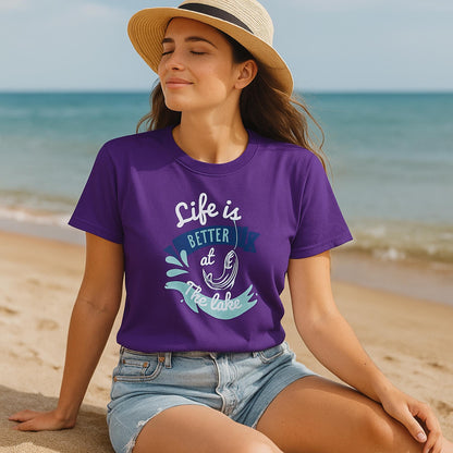 Relaxed woman sitting on a sandy beach wearing a purple t-shirt with a colorful graphic that says &#39;Life is Better at the Lake,&#39; featuring a fish hook, water splash, and banner design. She is also wearing a straw hat and denim shorts, with calm lake water and a clear sky in the background.