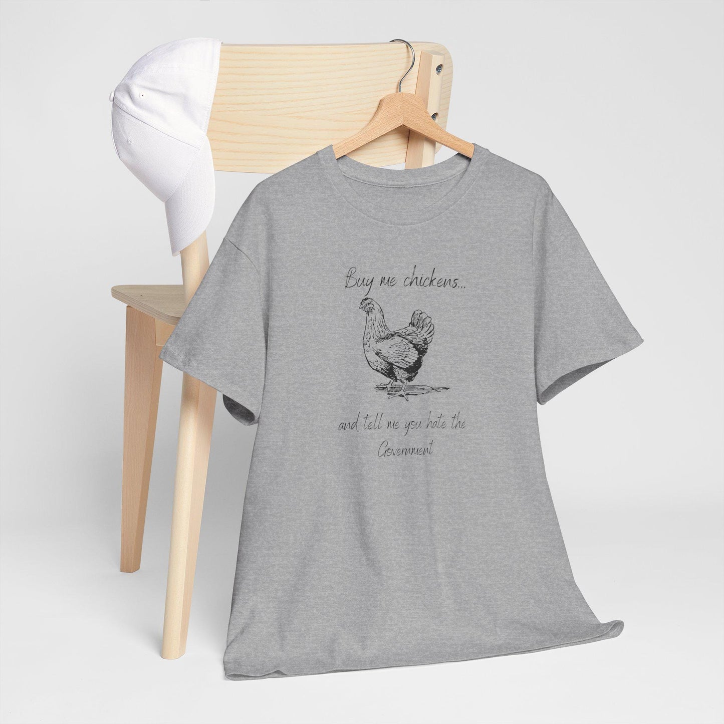 A heather gray T-shirt hangs on a wooden hanger placed over the back of a light wood chair. A white baseball cap rests on the chair. The T-shirt features a sketch-style illustration of a chicken and the humorous text: &quot;Buy me chickens... and tell me you hate the Government.&quot; The setup is minimal and styled for a clean, casual product display.
