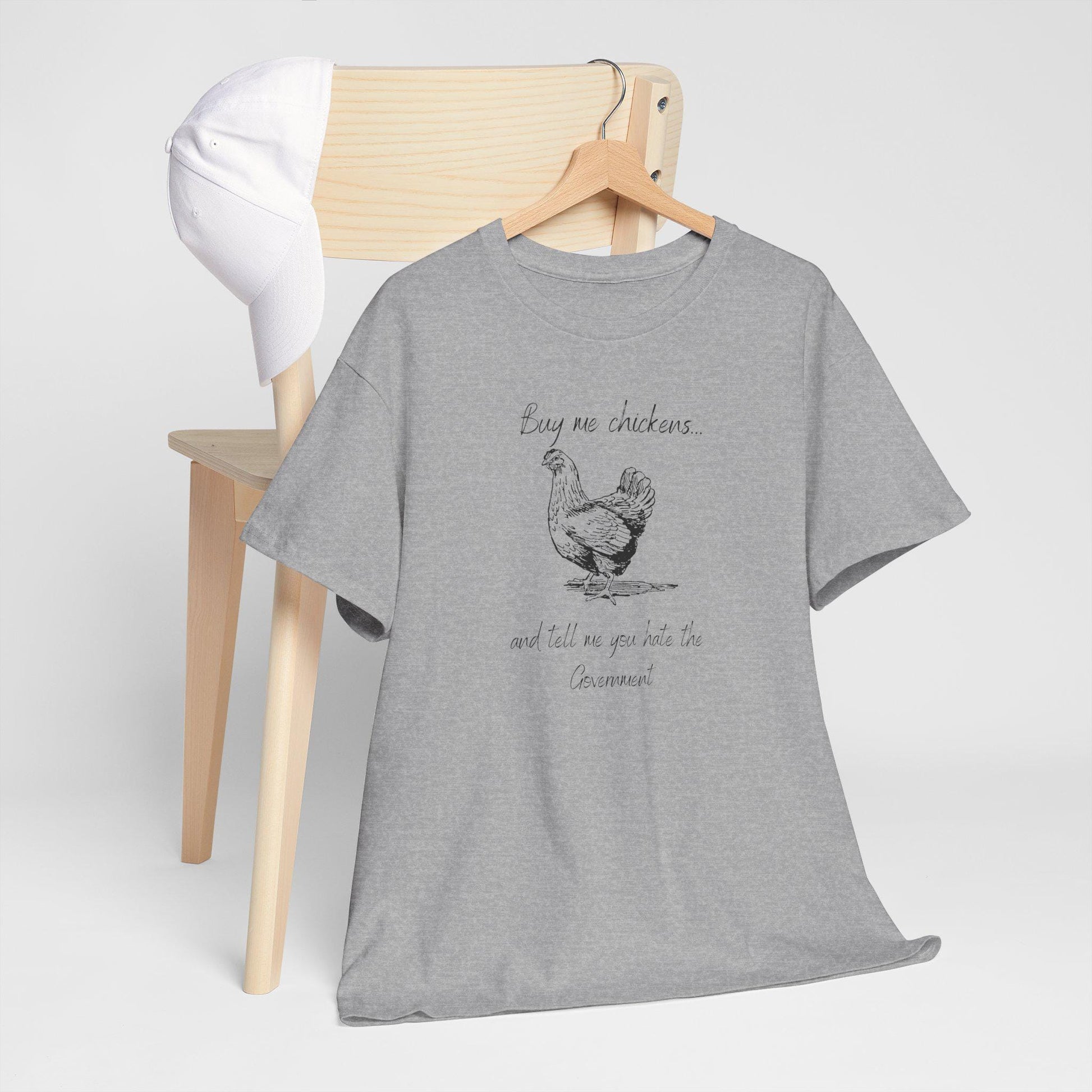 A heather gray T-shirt hangs on a wooden hanger placed over the back of a light wood chair. A white baseball cap rests on the chair. The T-shirt features a sketch-style illustration of a chicken and the humorous text: &quot;Buy me chickens... and tell me you hate the Government.&quot; The setup is minimal and styled for a clean, casual product display.