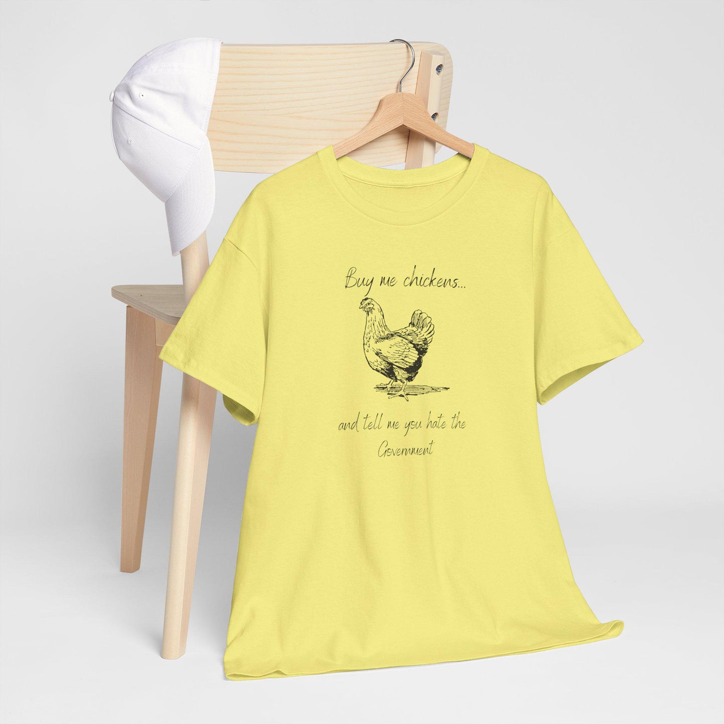A light yellow T-shirt is displayed on a wooden hanger draped over the back of a light wood chair. A white baseball cap rests on the chair. The T-shirt features a vintage-style illustration of a chicken along with the humorous phrase: &quot;Buy me chickens... and tell me you hate the Government.&quot; The setup is clean and minimal, emphasizing the shirt&#39;s design.