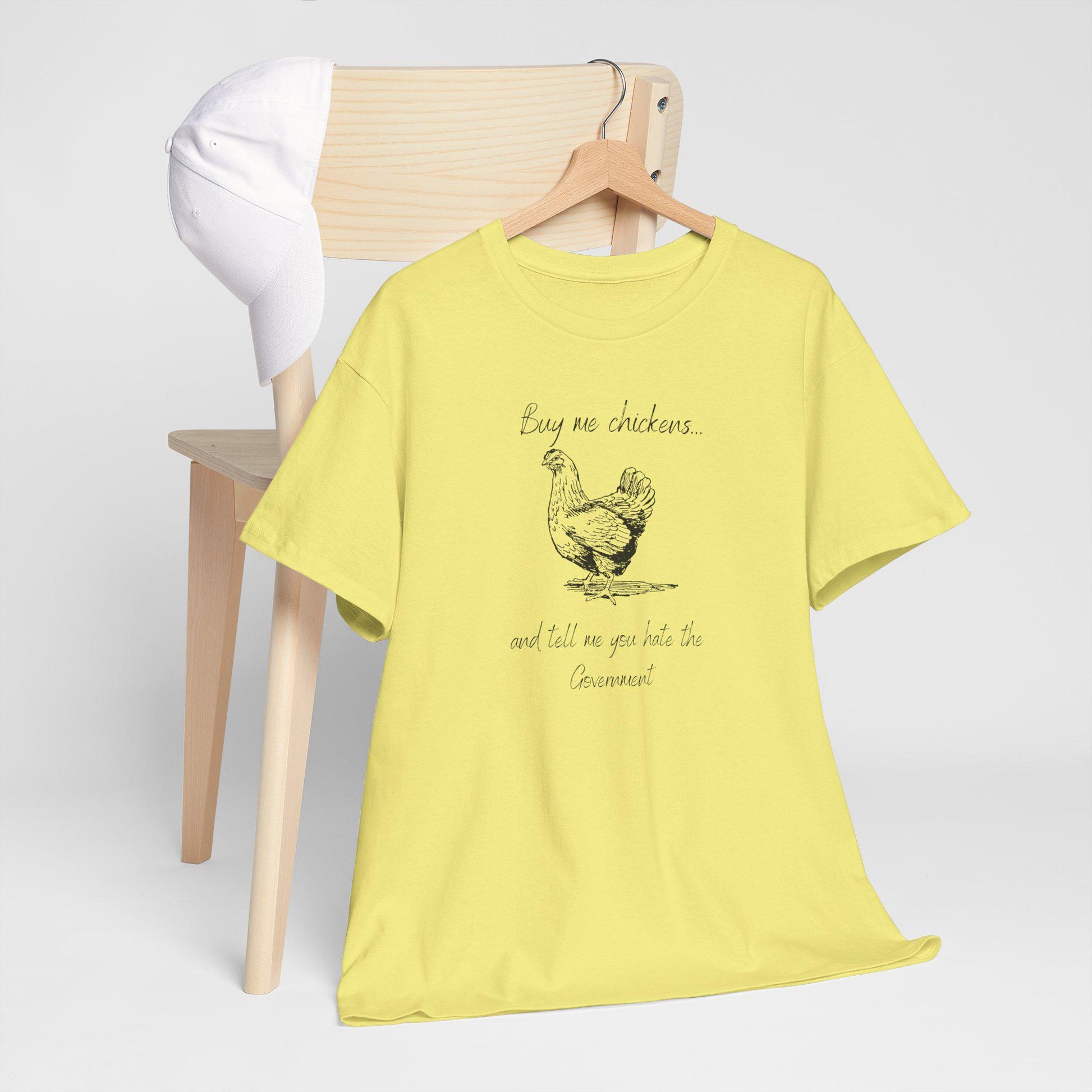 A light yellow T-shirt is displayed on a wooden hanger draped over the back of a light wood chair. A white baseball cap rests on the chair. The T-shirt features a vintage-style illustration of a chicken along with the humorous phrase: &quot;Buy me chickens... and tell me you hate the Government.&quot; The setup is clean and minimal, emphasizing the shirt&#39;s design.