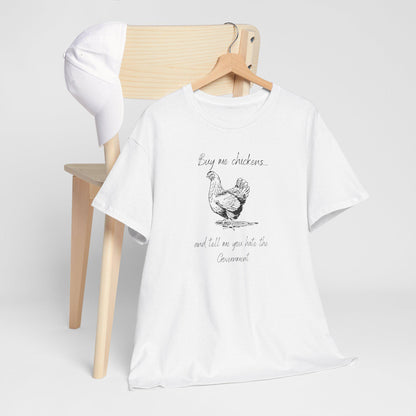 A white T-shirt hangs on a wooden hanger placed over the back of a light wood chair, with a white baseball cap resting on the chair. The T-shirt features a black sketch-style illustration of a chicken and the humorous phrase: &quot;Buy me chickens... and tell me you hate the Government.&quot; The display is minimal and clean, highlighting the shirt&#39;s design.