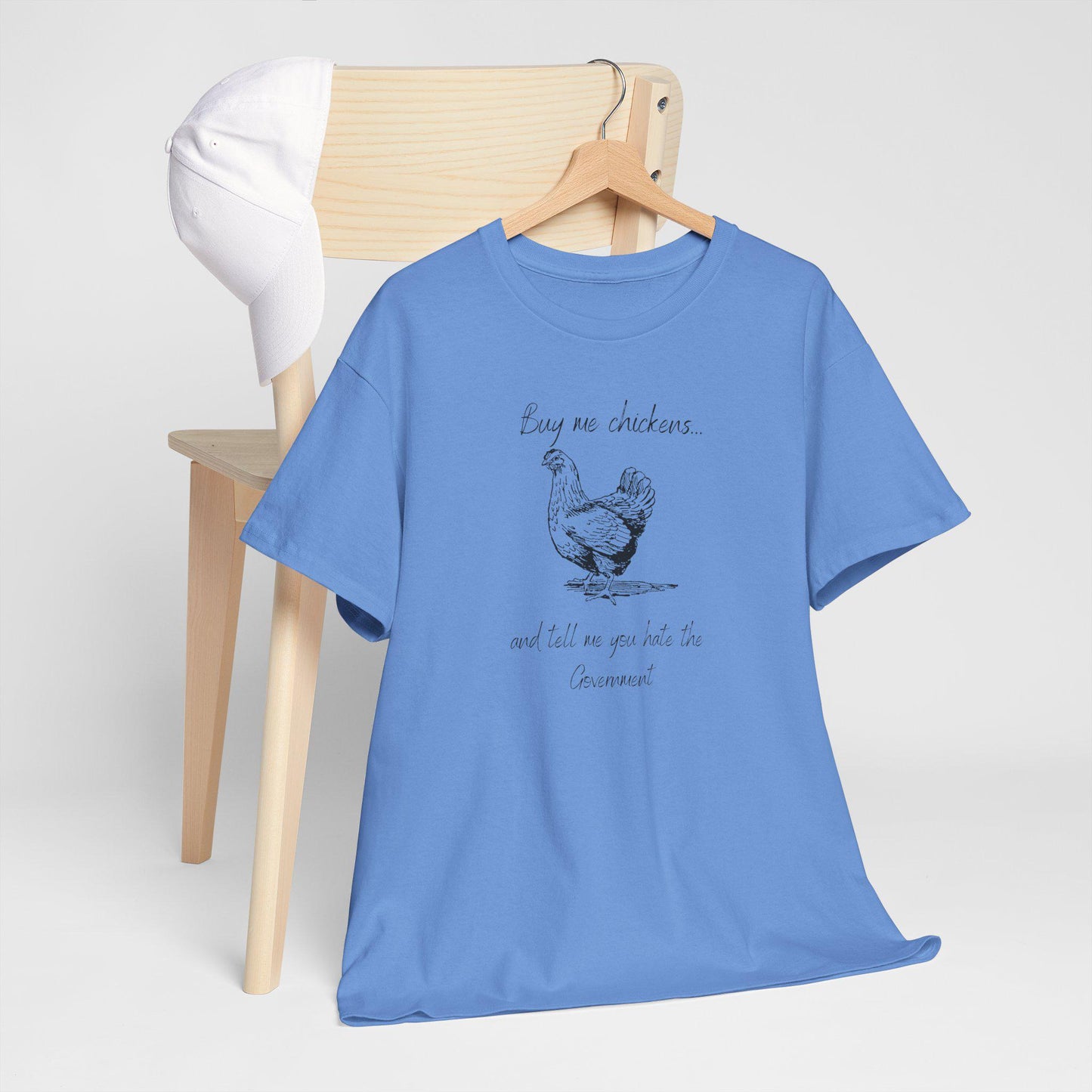 A light blue T-shirt hangs on a wooden hanger draped over the back of a light wood chair, with a white baseball cap resting on the chair. The T-shirt features a hand-drawn style illustration of a chicken and the humorous text: &quot;Buy me chickens... and tell me you hate the Government.&quot; The clean, minimal display highlights the shirt’s casual design and witty message.
