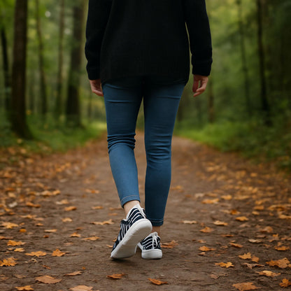 Woman walking on a leaf-covered forest trail wearing blue skinny jeans, a black sweater, and black-and-white swirl patterned sneakers. Peaceful autumn nature walk, perfect for promoting casual fall fashion, stylish walking shoes, and outdoor lifestyle content.