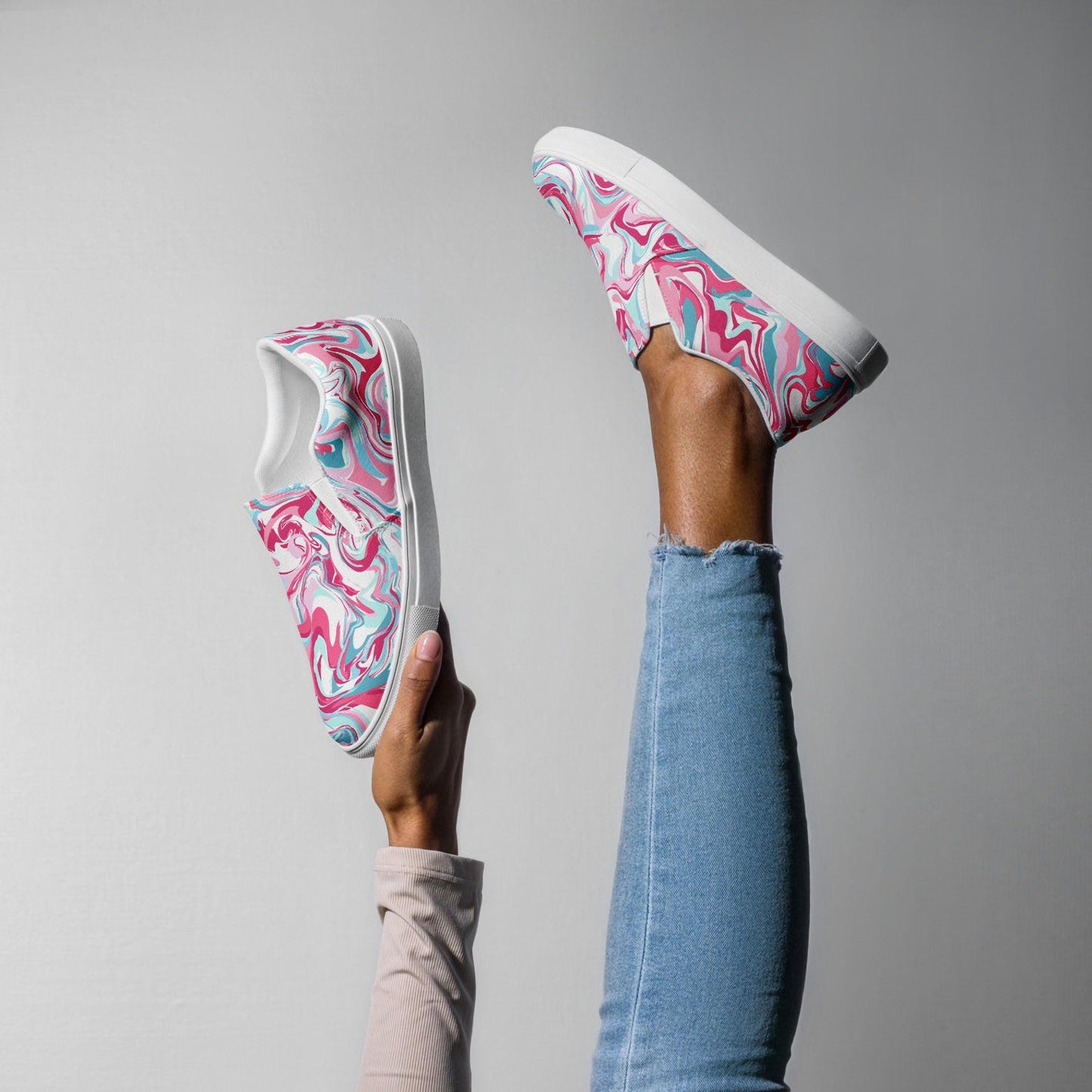 A person poses creatively against a plain background with one leg raised in the air wearing a colorful slip-on sneaker featuring a pink, teal, and white marbled swirl pattern. Their other hand holds the matching sneaker upright. The person is wearing light blue jeans and a light beige long-sleeve top.