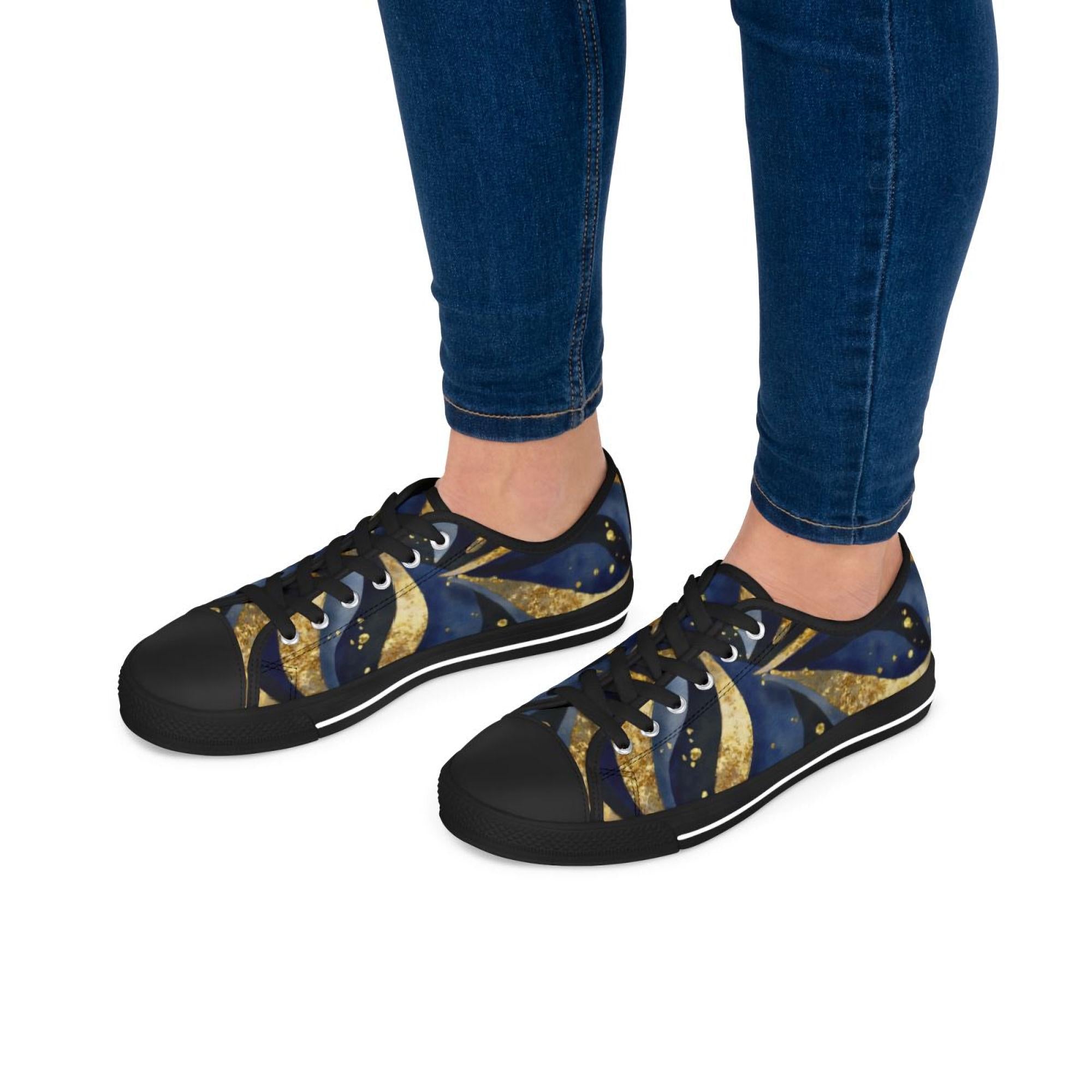 A person wearing low-top canvas sneakers with a celestial-themed design featuring swirling deep blue and gold patterns resembling stars and comets. The shoes have black soles, black laces, and black toe caps, adding contrast to the vibrant print. The person is dressed in fitted blue jeans, and the background is plain white, keeping the focus on the shoes.