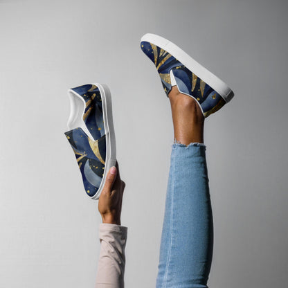 A person poses playfully against a plain gray background, holding one slip-on sneaker in their hand while wearing the other on a raised leg. The sneakers feature a bold celestial-themed design with swirling deep blue and gold patterns, and have clean white soles. The person is dressed in light blue skinny jeans and a beige long-sleeve top, showcasing the artistic footwear in a fun and creative way.