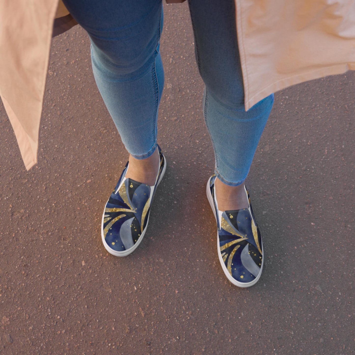 A top-down view of a person wearing slip-on sneakers with a celestial-themed design in deep blue and gold, featuring swirling patterns and small star-like dots. The person is dressed in light blue skinny jeans and a beige coat, standing on a textured reddish pavement. The warm lighting suggests a sunrise or sunset setting.