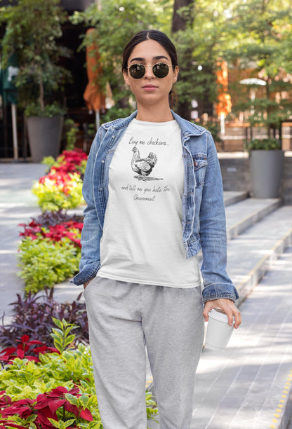 A woman stands confidently on a city sidewalk holding a coffee cup. She is wearing sunglasses, a denim jacket over a white T-shirt, and gray sweatpants. Her T-shirt features a sketch of a chicken and the humorous phrase: &quot;Buy me chickens... and tell me you hate the Government.&quot;