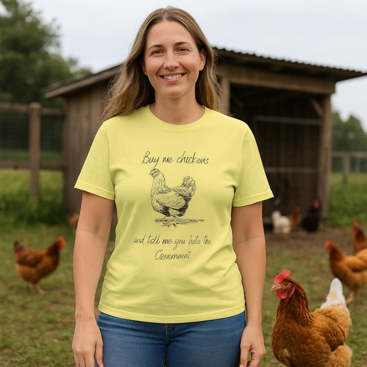 A woman stands smiling in front of a chicken coop surrounded by brown hens. She is wearing a light yellow T-shirt featuring a hand-drawn illustration of a chicken and the humorous text: &quot;Buy me chickens... and tell me you hate the Government.&quot;