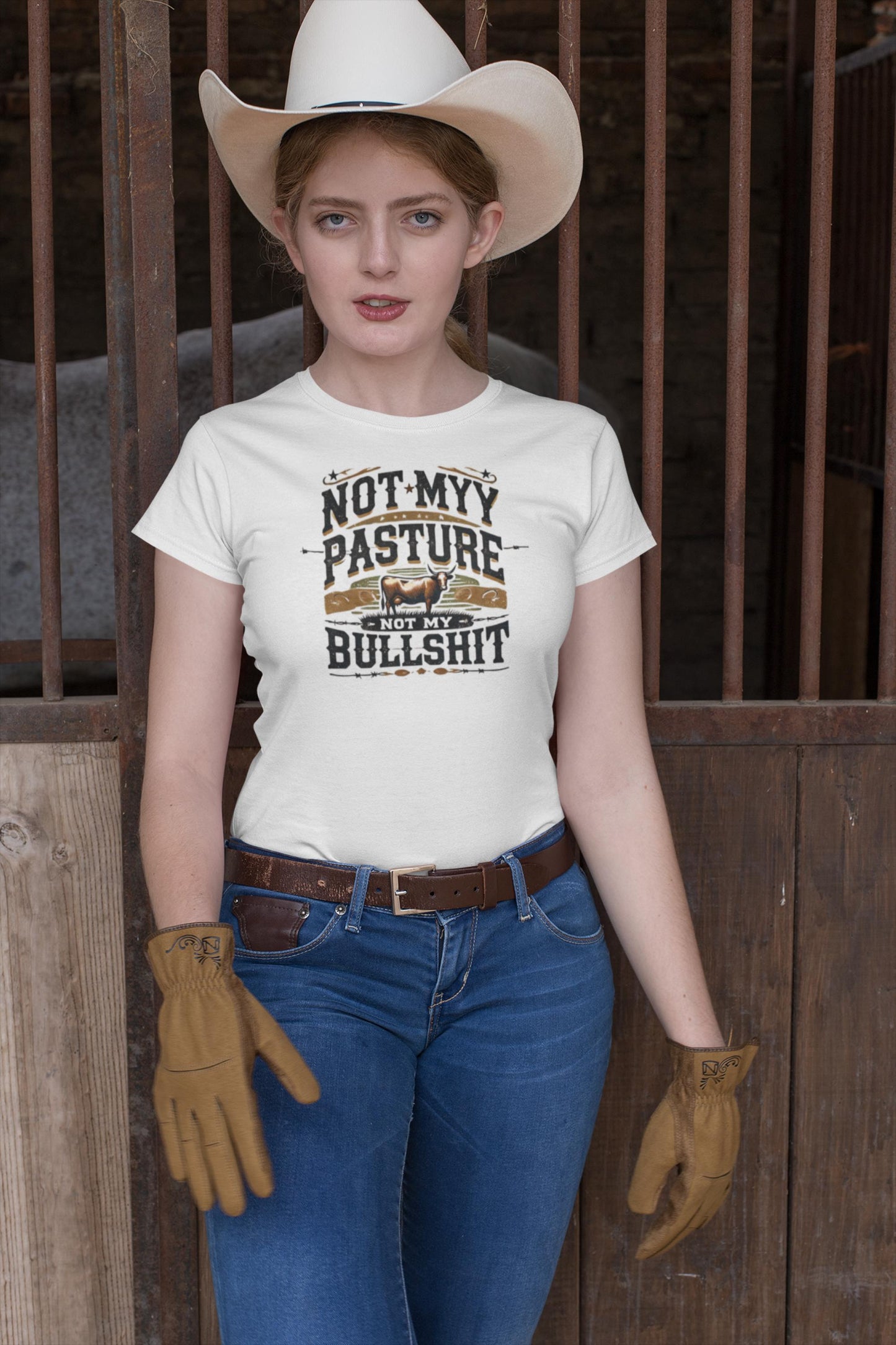A woman wearing a white cowboy hat, brown work gloves, and blue jeans stands in front of a rustic barn gate. She is dressed in a fitted white T-shirt with a bold, western-style graphic that includes a cow and reads: &quot;Not my pasture, not my bullshit.&quot; Her stance and expression convey confidence and a no-nonsense attitude in a country setting.