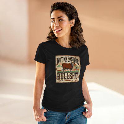 A woman stands indoors in a warmly lit space, smiling and wearing a black T-shirt paired with blue jeans. The shirt features a vintage-style graphic of a brown cow in a western landscape with the bold, humorous text: &quot;Not my pasture, not my bullshit.&quot; She poses casually with her hands resting in her pockets.