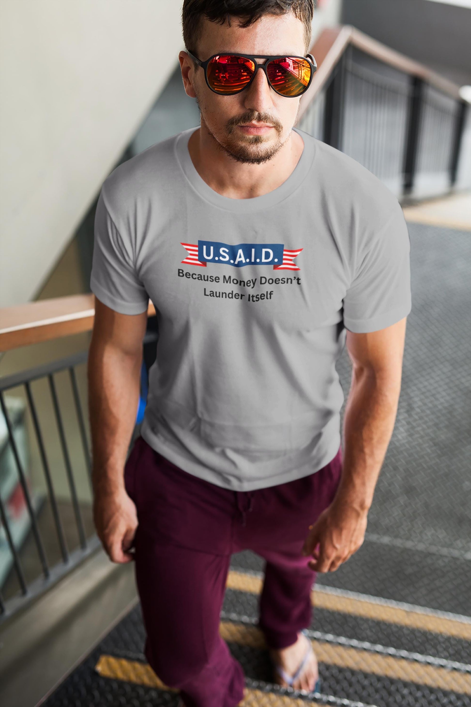 A man walks up a staircase wearing red mirrored sunglasses, burgundy joggers, flip-flops, and a light gray T-shirt. The shirt features a satirical political design with a navy blue banner reading &quot;U.S.A.I.D.&quot; flanked by red and white striped ribbons. Below it, black text states: &quot;Because Money Doesn’t Launder Itself.&quot; The man has a confident expression, and the setting appears to be a modern indoor public space.