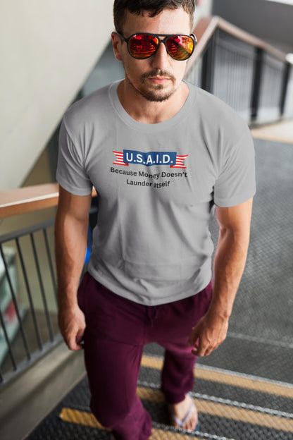 A man walks up a staircase wearing red mirrored sunglasses, burgundy joggers, flip-flops, and a light gray T-shirt. The shirt features a satirical political design with a navy blue banner reading &quot;U.S.A.I.D.&quot; flanked by red and white striped ribbons. Below it, black text states: &quot;Because Money Doesn’t Launder Itself.&quot; The man has a confident expression, and the setting appears to be a modern indoor public space.