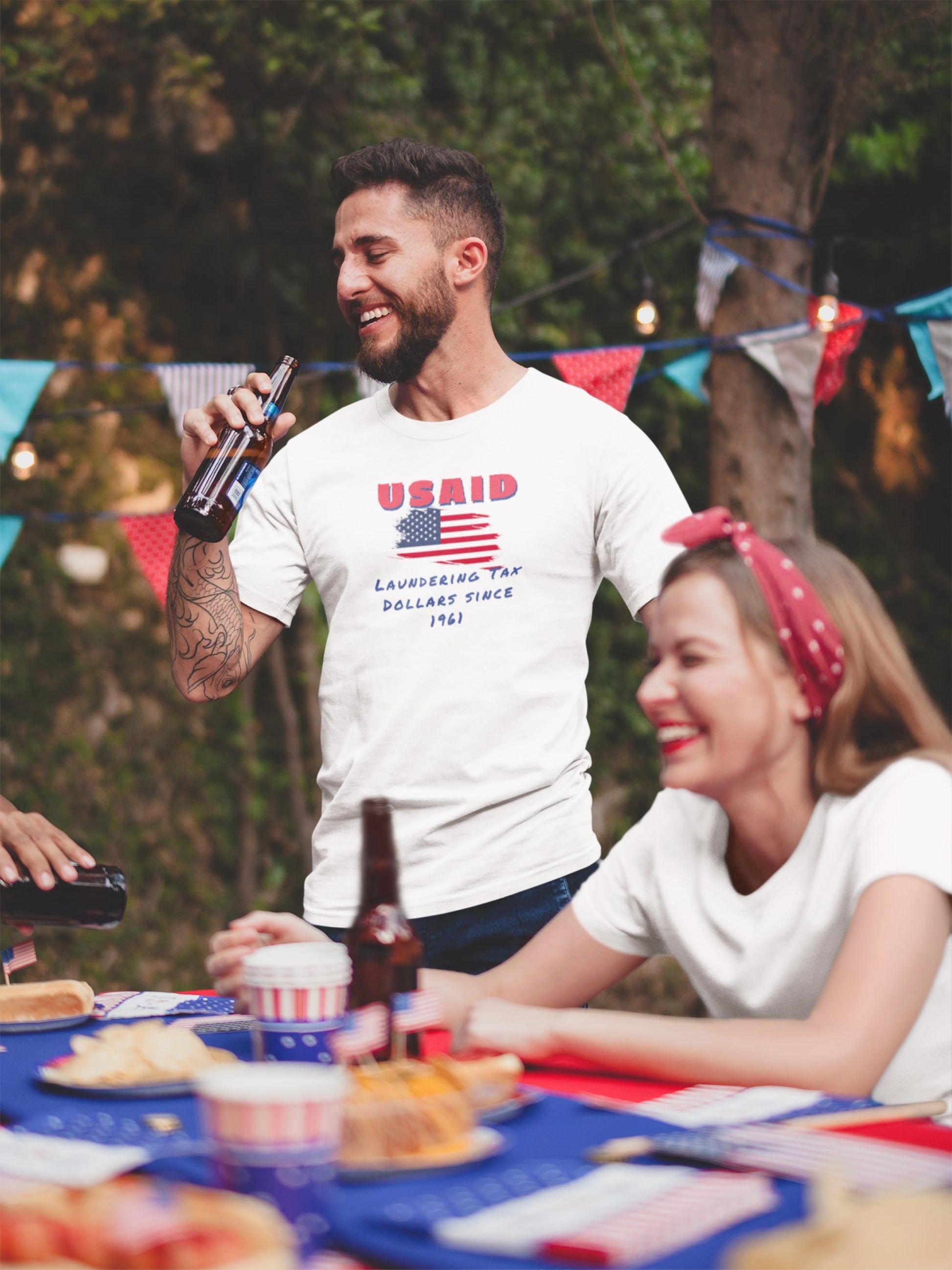 A man laughs while holding a beer at an outdoor party decorated with red, white, and blue bunting and patriotic tableware. He is wearing a white T-shirt with a distressed American flag graphic and the text: &quot;USAID&quot; in bold red letters above, and &quot;Laundering Tax Dollars Since 1961&quot; in blue handwritten-style text below. A woman next to him, also smiling, is seated at a table filled with festive food and drinks.
