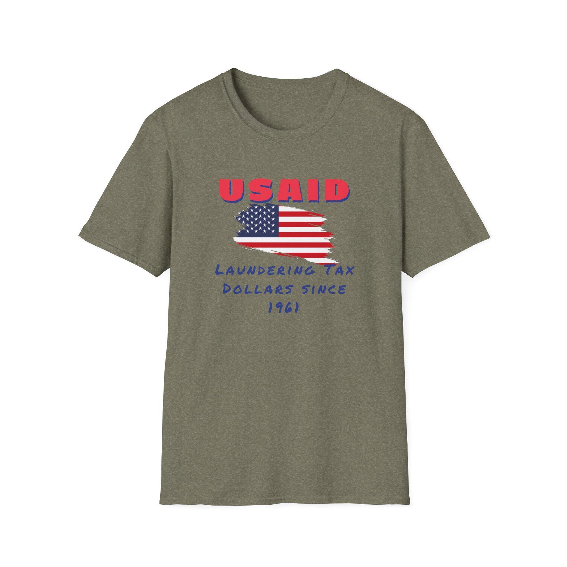 An army green T-shirt is laid flat against a white background. The shirt features a distressed American flag graphic in the center. Above the flag, bold red text reads &quot;USAID&quot; in all caps. Below, blue handwritten-style text says: &quot;Laundering Tax Dollars Since 1961.&quot; The design delivers a bold, satirical political message with a rugged protest-inspired style.