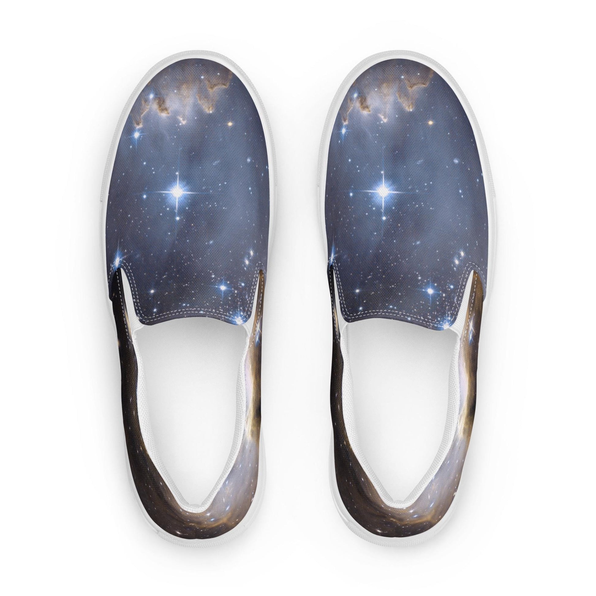 Top-down view of a pair of slip-on sneakers featuring a stunning galaxy print with stars, nebulae, and cosmic clouds in deep blues and subtle gold tones. The shoes have white elastic side panels and white soles, displayed against a clean white background.