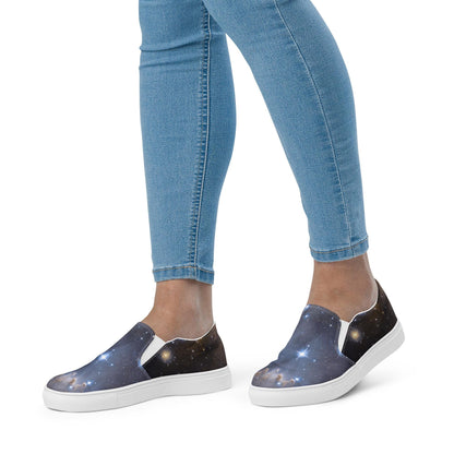 Close-up view of a person wearing galaxy-themed slip-on sneakers featuring a vivid outer space print with stars and nebulae. The shoes have white elastic side panels and white soles, paired with light blue skinny jeans, against a plain white background.