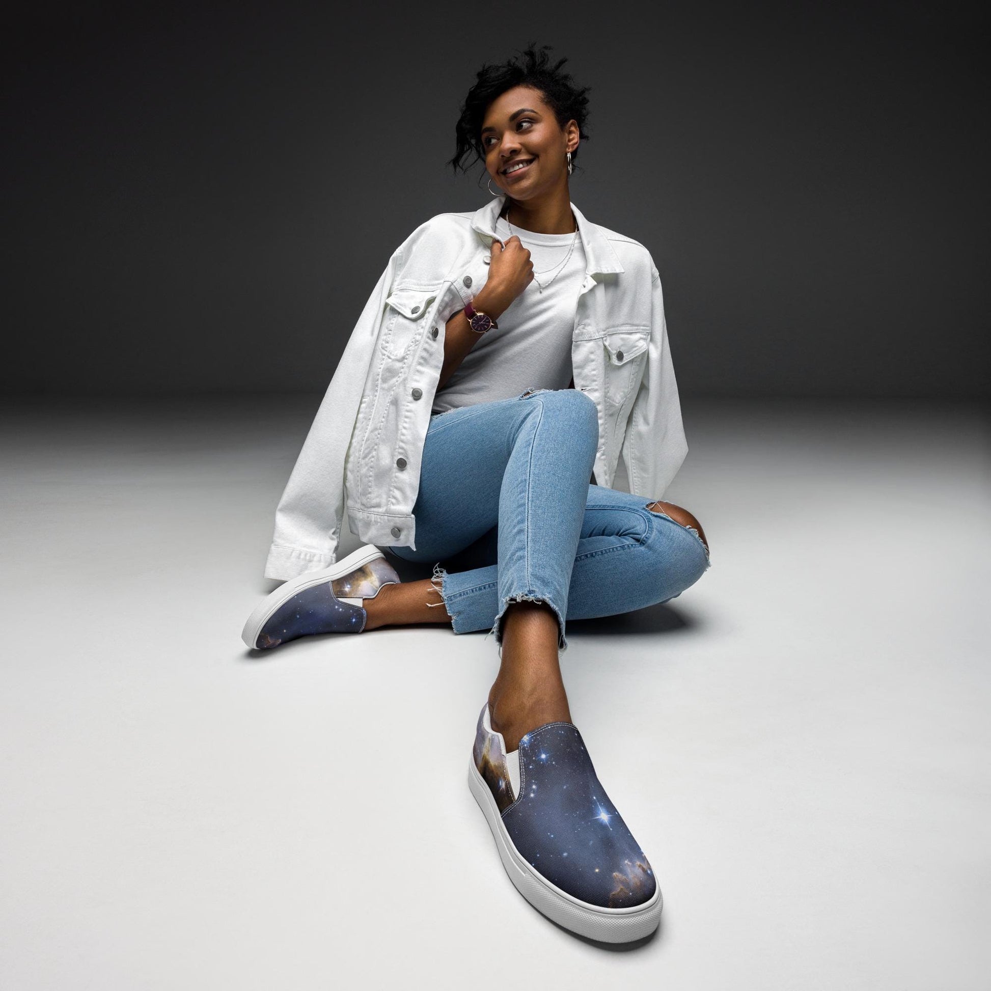 Stylish woman sitting on the floor in a minimalist studio setting, wearing a white denim jacket, white t-shirt, ripped light blue jeans, and galaxy-themed slip-on sneakers with white soles. She is smiling and posed casually with one leg crossed, showcasing her cosmic-inspired footwear.