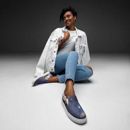 Stylish woman sitting on the floor in a minimalist studio setting, wearing a white denim jacket, white t-shirt, ripped light blue jeans, and galaxy-themed slip-on sneakers with white soles. She is smiling and posed casually with one leg crossed, showcasing her cosmic-inspired footwear.