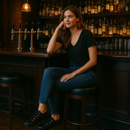 A young woman with long brown hair is sitting on a barstool at a dimly lit bar. She is wearing a black V-neck T-shirt, dark blue jeans, and black sneakers with gold eyelets. She rests her head on one hand and smiles slightly, appearing relaxed. The background features shelves filled with liquor bottles and a row of beer taps on the bar counter.