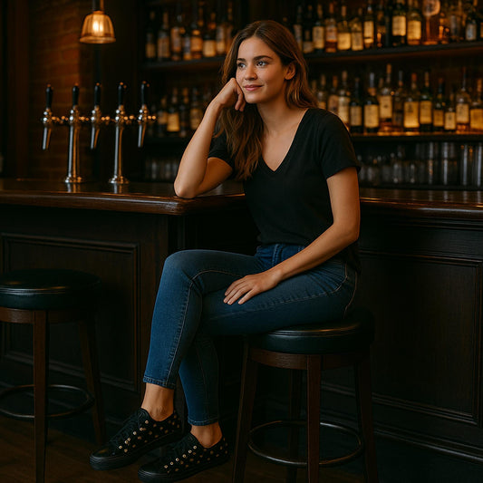 A young woman with long brown hair is sitting on a barstool at a dimly lit bar. She is wearing a black V-neck T-shirt, dark blue jeans, and black sneakers with gold eyelets. She rests her head on one hand and smiles slightly, appearing relaxed. The background features shelves filled with liquor bottles and a row of beer taps on the bar counter.