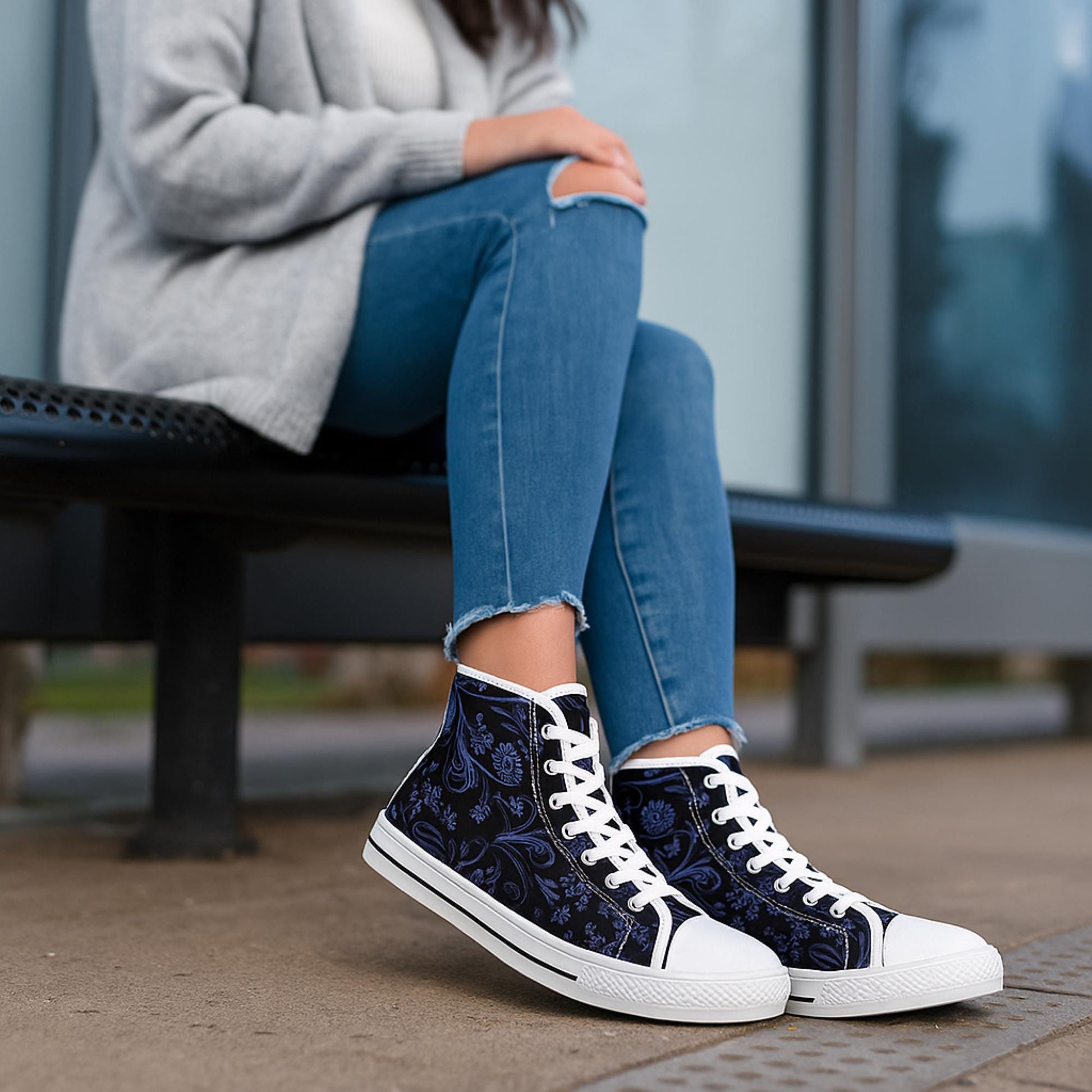 A woman is seated on a bench wearing light blue skinny jeans with frayed hems and a light gray sweater. She is wearing a pair of navy blue high-top sneakers with white soles, toe caps, and laces. The sneakers feature a delicate floral and paisley pattern in lighter blue tones. The setting appears to be an outdoor urban area near a glass building.