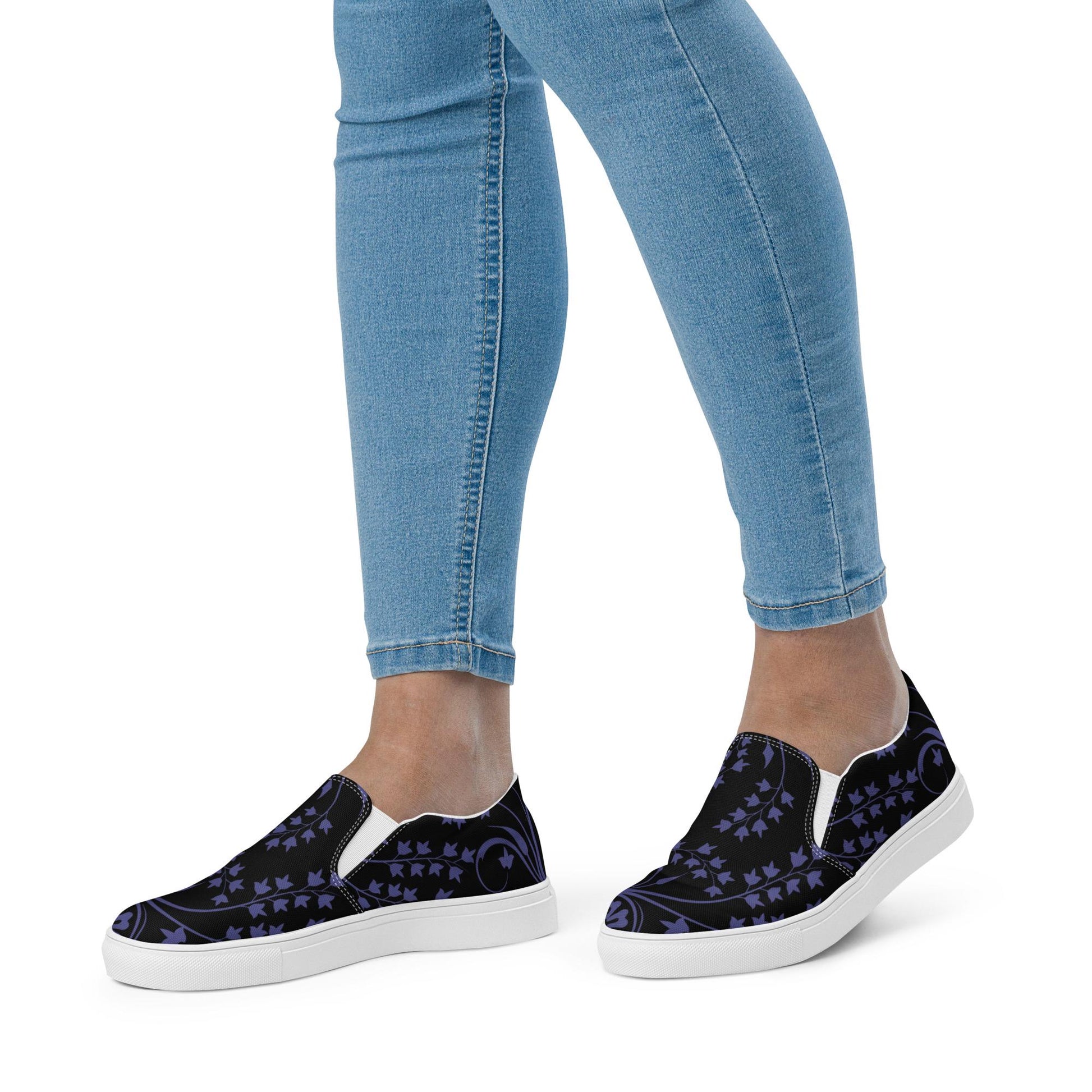 Person wearing slip-on sneakers with a black base and dark purple ivy vine design, paired with light blue skinny jeans. The shoes feature white midsoles and elastic side panels for easy wear, creating a comfortable and stylish everyday look.