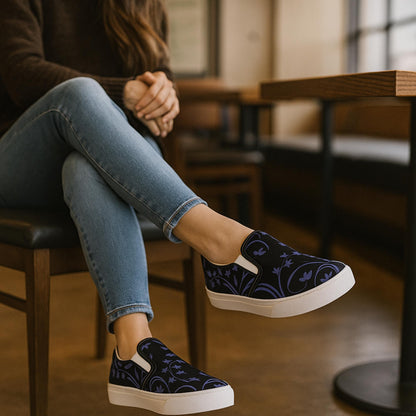 Person sitting in a cozy café wearing slip-on sneakers with a black background and dark purple floral vine design. The shoes are paired with light blue skinny jeans and a dark sweater, creating a relaxed, stylish look perfect for a casual outing.