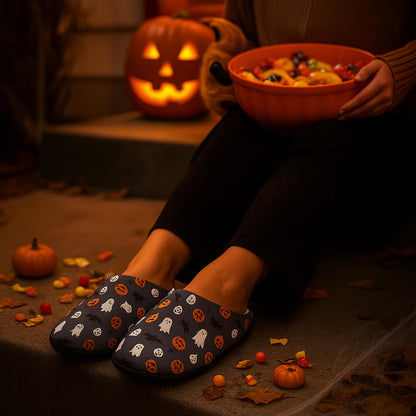 Person sitting on a porch at night wearing Halloween-themed slippers decorated with ghosts, pumpkins, and bats. They are holding a large orange bowl filled with colorful candy, surrounded by small pumpkins, scattered candy pieces, and autumn leaves. A glowing carved jack-o&#39;-lantern adds a festive Halloween atmosphere.