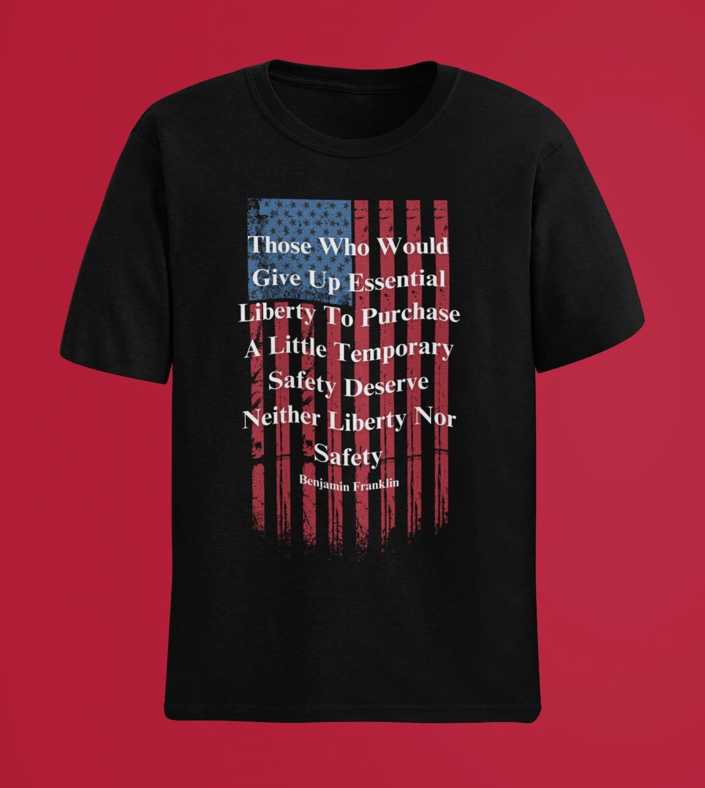 A black T-shirt is displayed against a solid red background. The shirt features a distressed American flag graphic in faded red and blue, overlaid with a bold white quote by Benjamin Franklin that reads: “Those Who Would Give Up Essential Liberty To Purchase A Little Temporary Safety Deserve Neither Liberty Nor Safety.” The clean and simple backdrop makes the patriotic design stand out clearly.
