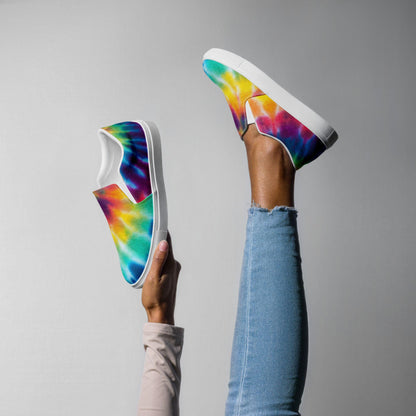 Woman playfully showcasing rainbow tie-dye slip-on sneakers with legs raised against a light gray background. The vibrant canvas shoes feature a bold mix of blue, green, yellow, orange, red, and purple in a dreamy watercolor swirl design. Styled with a clean white rubber sole and easy slip-on fit, these sneakers pair perfectly with light-wash skinny jeans.