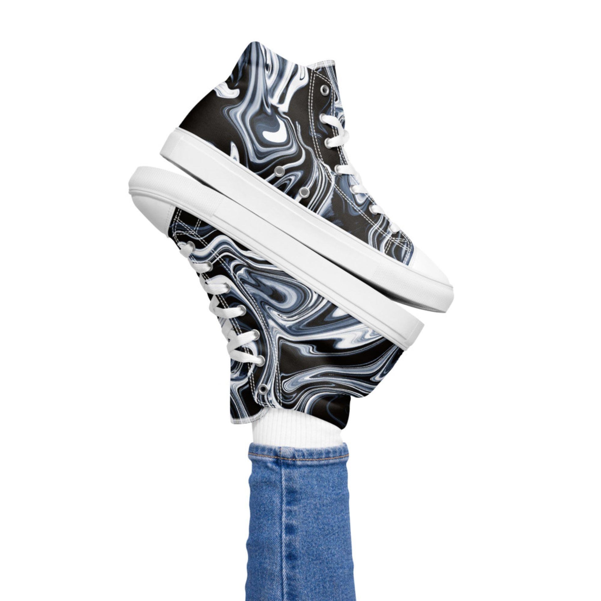 Playful image of a person lifting both legs while wearing high-top sneakers with a black, white, and blue marble swirl design. The shoes feature white laces and soles, paired with white socks and blue skinny jeans, set against a plain white background.