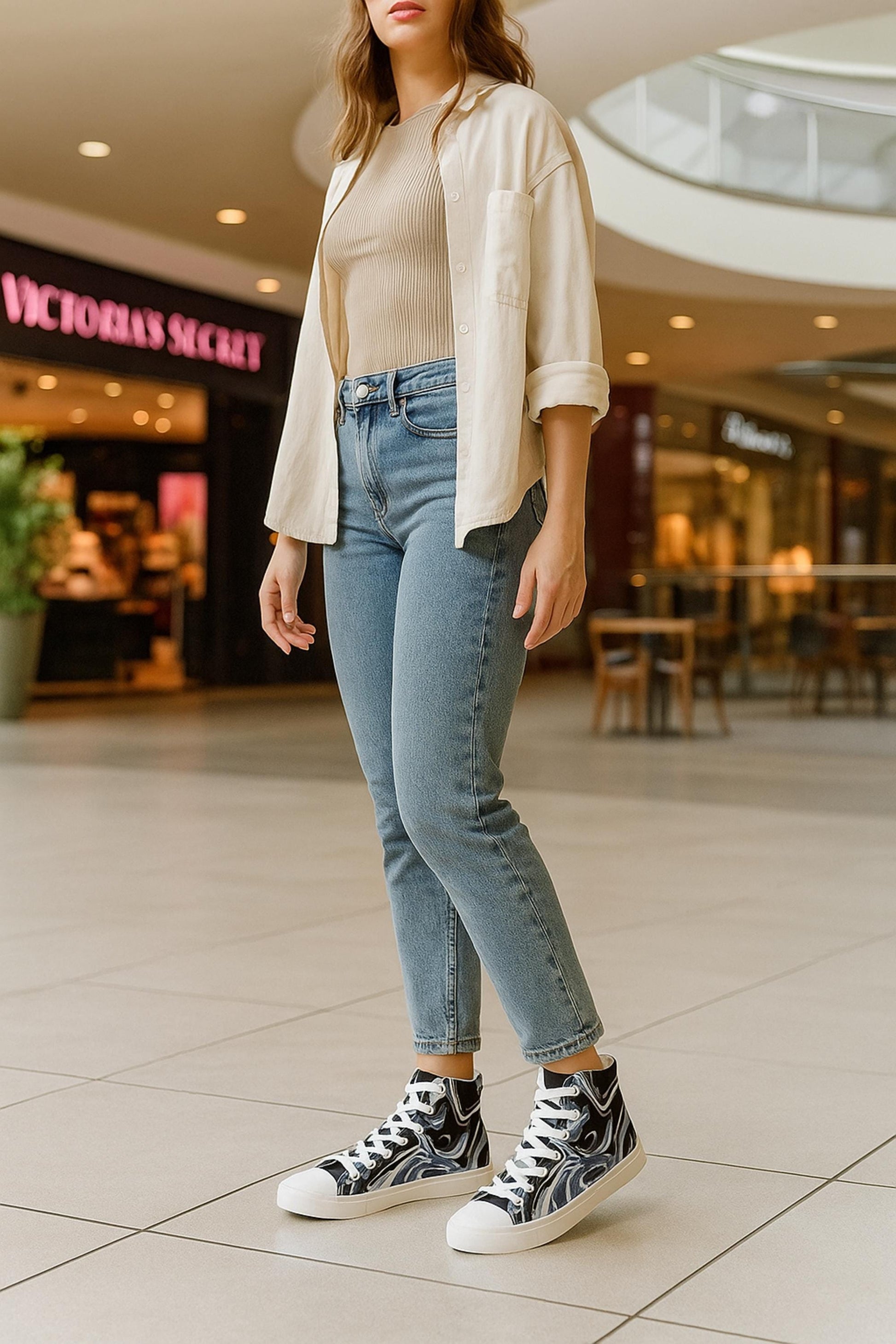 A woman standing in a shopping mall wearing light blue high-waisted jeans, a beige ribbed tank top, and a cream button-up shirt with rolled sleeves. She is wearing high-top sneakers with a black and white abstract swirl design, white laces, and white toe caps. The image focuses on her outfit from the shoulders down, highlighting the stylish shoes against the modern indoor setting.