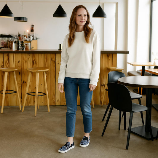 Woman wearing black and white abstract swirl canvas slip-on sneakers, blue jeans, and a white sweatshirt standing in a modern café with wood and black décor.